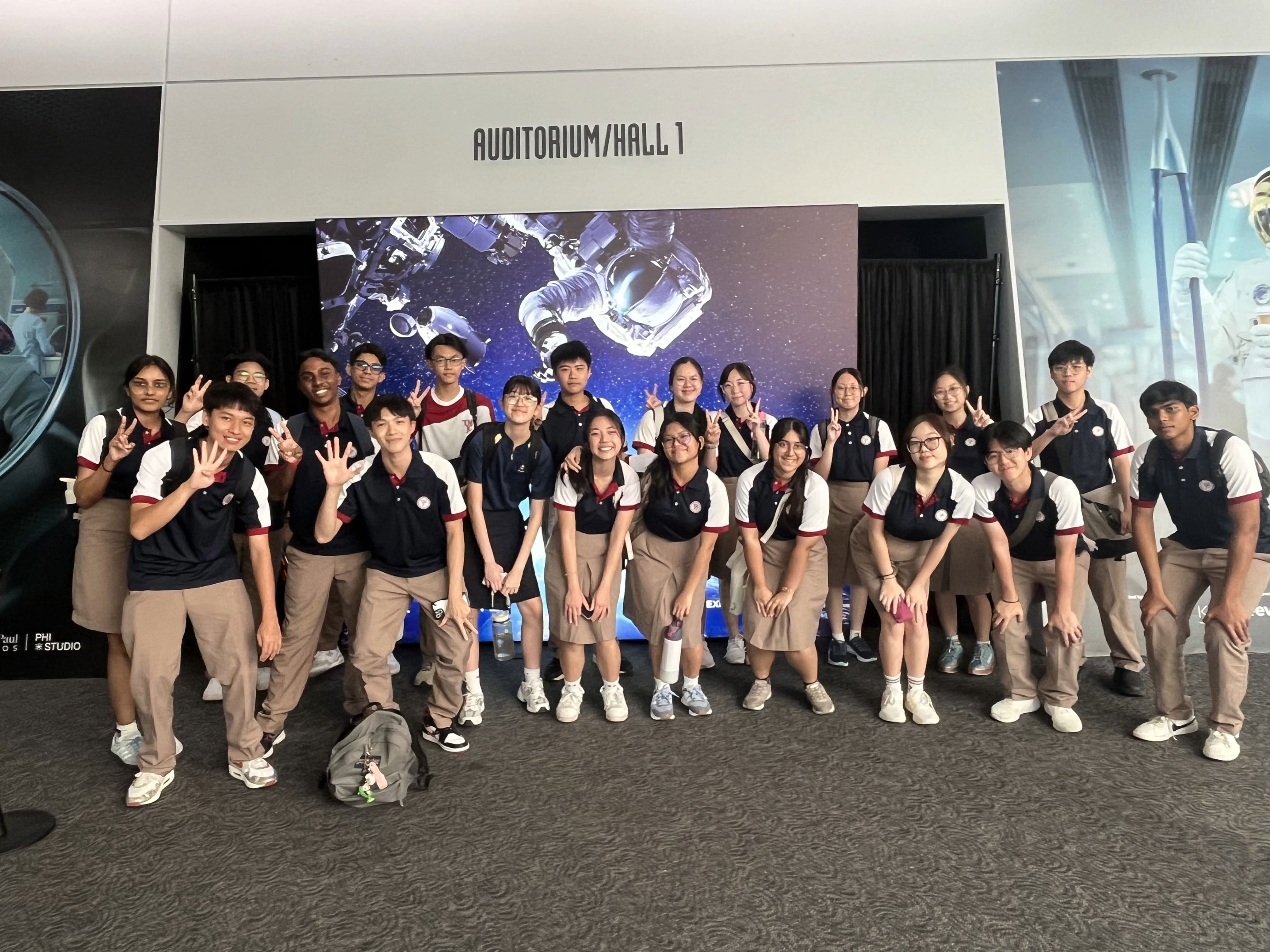 A large group of students in school uniforms poses energetically in front of a space-themed backdrop at the Science Centre auditorium, many flashing peace signs and smiling.